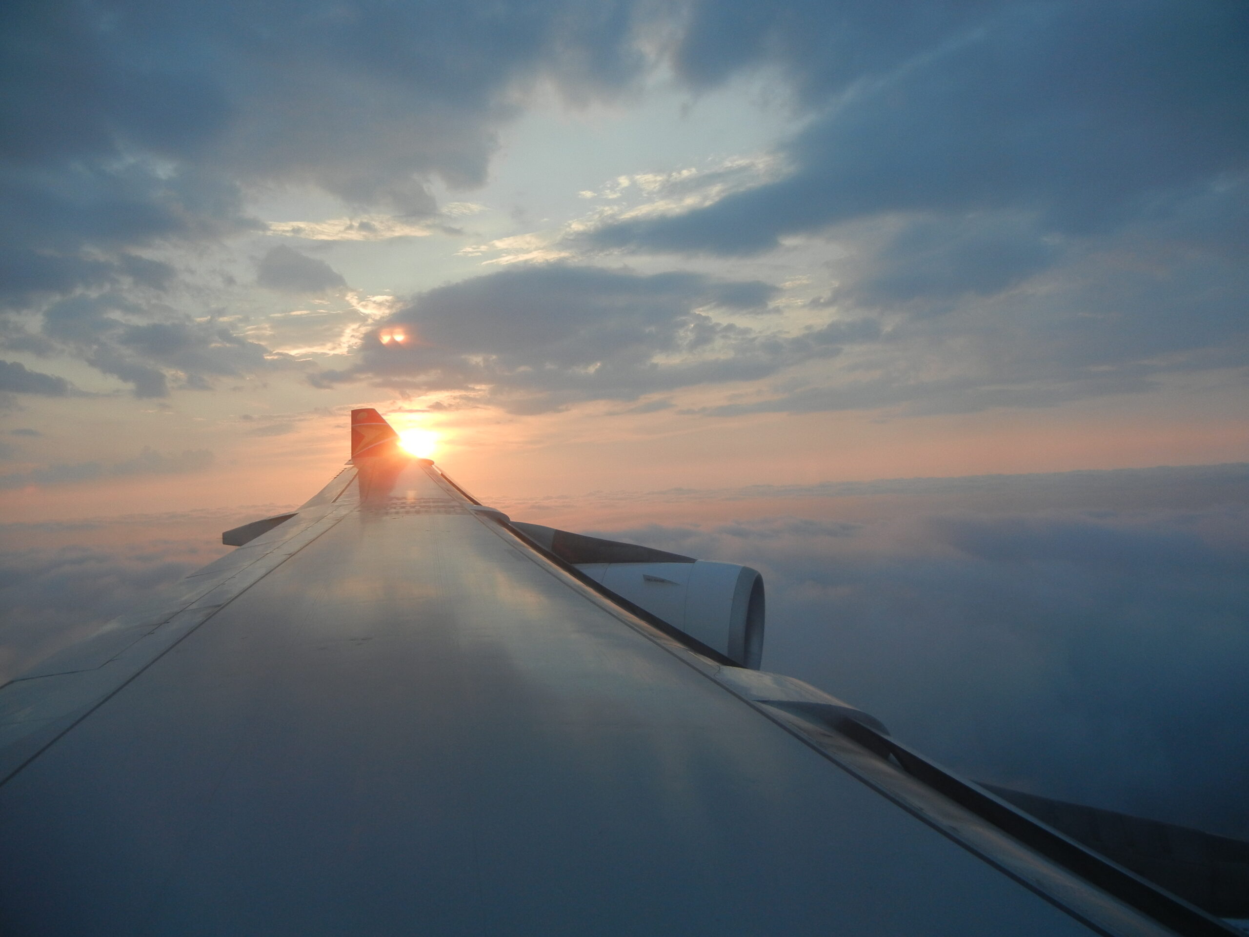 View from an airplane window showing the wing and engine with the sun setting above a layer of clouds.