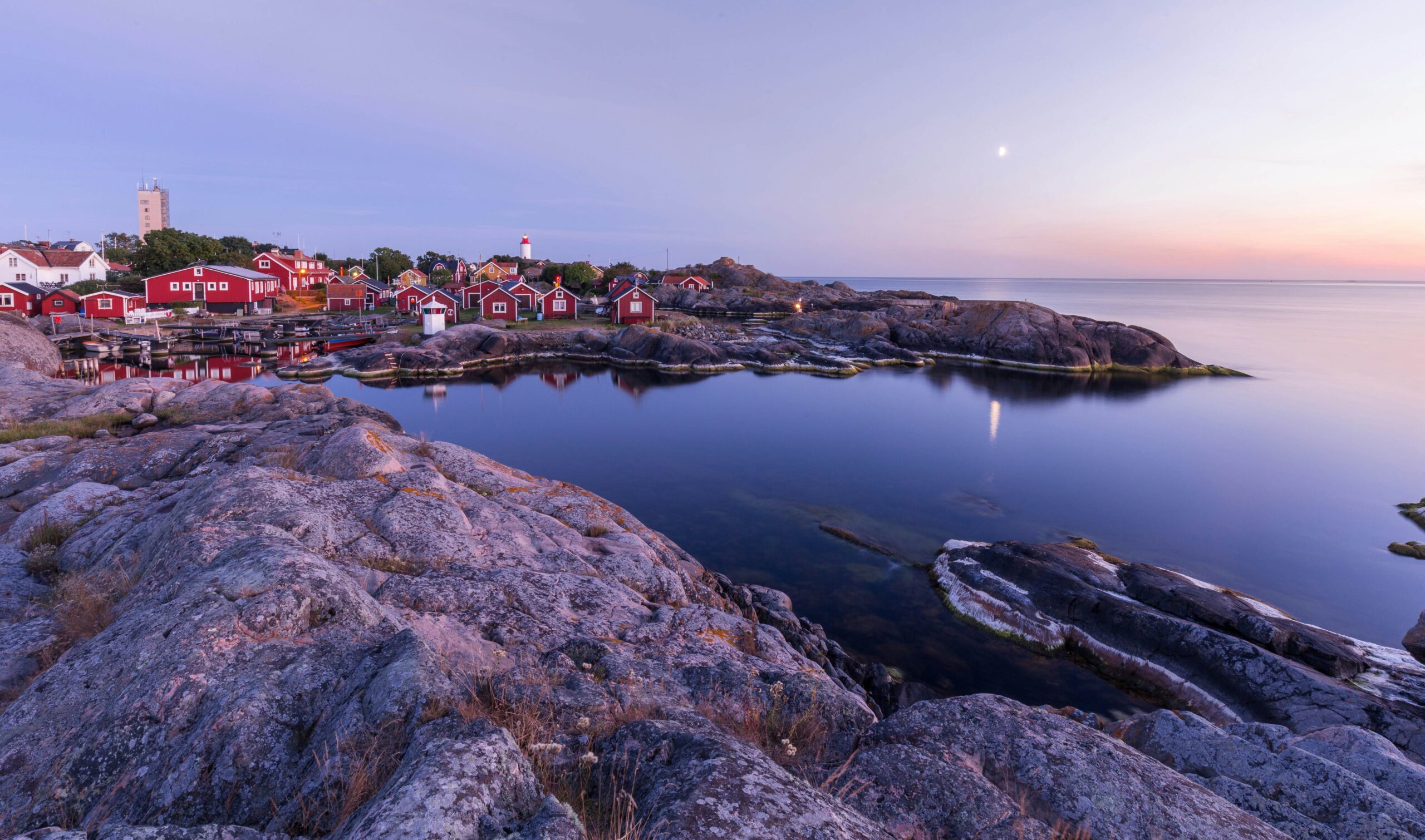 A rocky shoreline with a calm bay, red houses, a lighthouse, and a distant moon under a pastel sky at sunset.