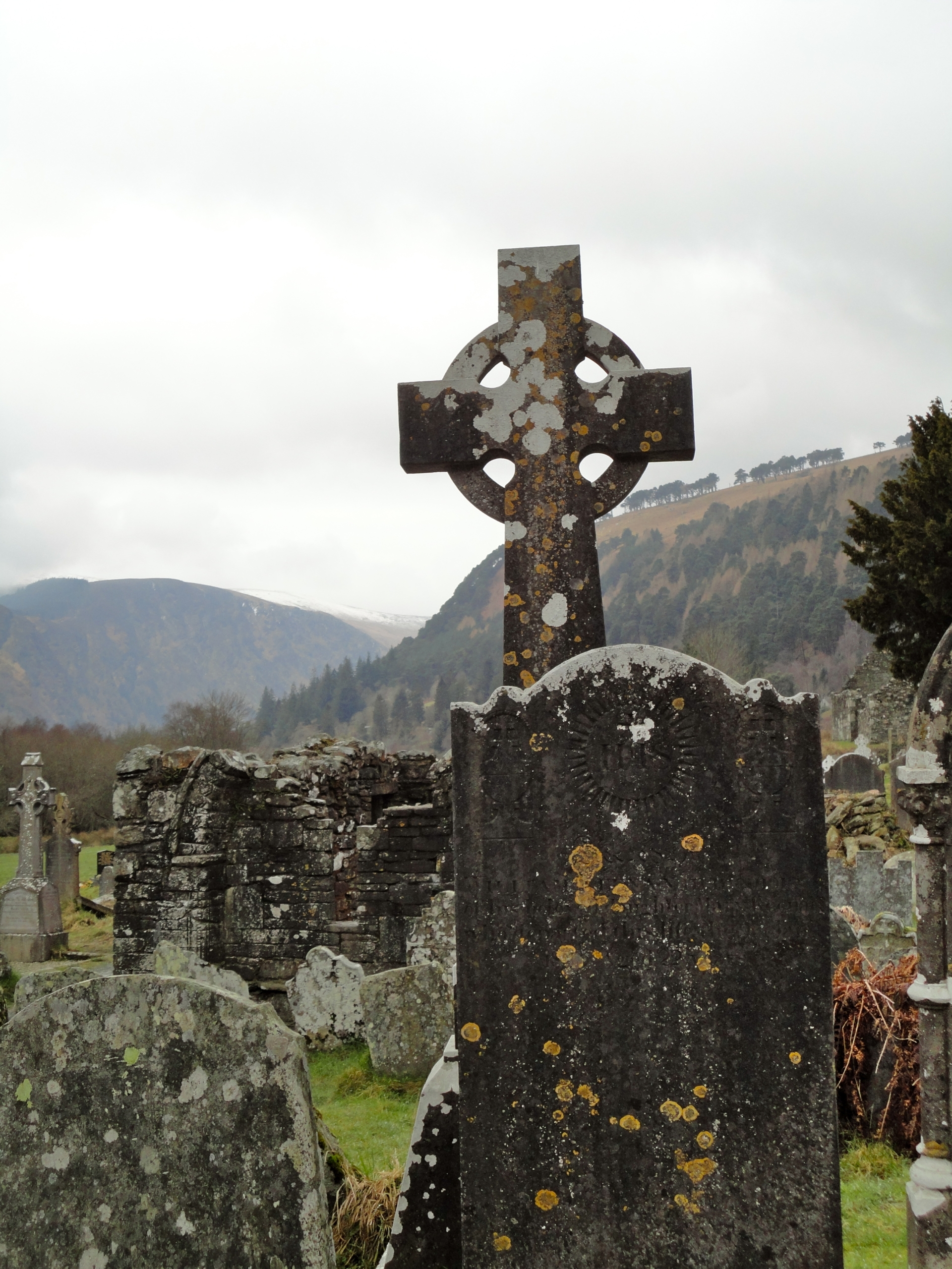 Weathered Celtic cross gravestone stands among other old tombstones in a cemetery, with mountains and cloudy sky in the background.