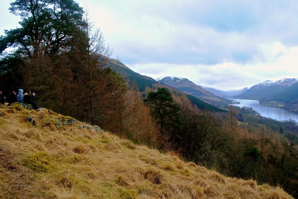 A group of people stand on a grassy hillside overlooking a forested valley, a lake, and snow-capped mountains under a cloudy sky.