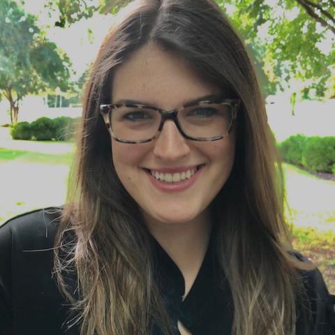 Woman with long brown hair and glasses smiling outdoors in a park-like setting with trees and greenery in the background.
