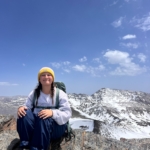 Person wearing a yellow beanie and backpack sits on a rocky mountain ledge with snow-covered peaks and partly cloudy sky in the background.
