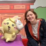 A person sits next to a large yellow cartoon character holding a mask, with a colorful illustrated background behind them.