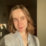 A person with short, wavy hair and light skin smiles at the camera in warm sunlight, wearing a light-colored, collared shirt.