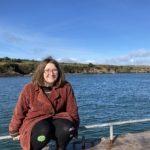 A person wearing glasses and a rust-colored coat sits on a railing by a body of water, with grassy hills and a blue sky in the background.
