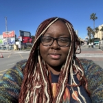 A person with long, braided hair and glasses is outdoors on a sunny day, standing in front of a street with billboards and palm trees visible in the background.
