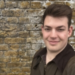 A person with short brown hair is smiling and taking a selfie in front of a textured brick wall.