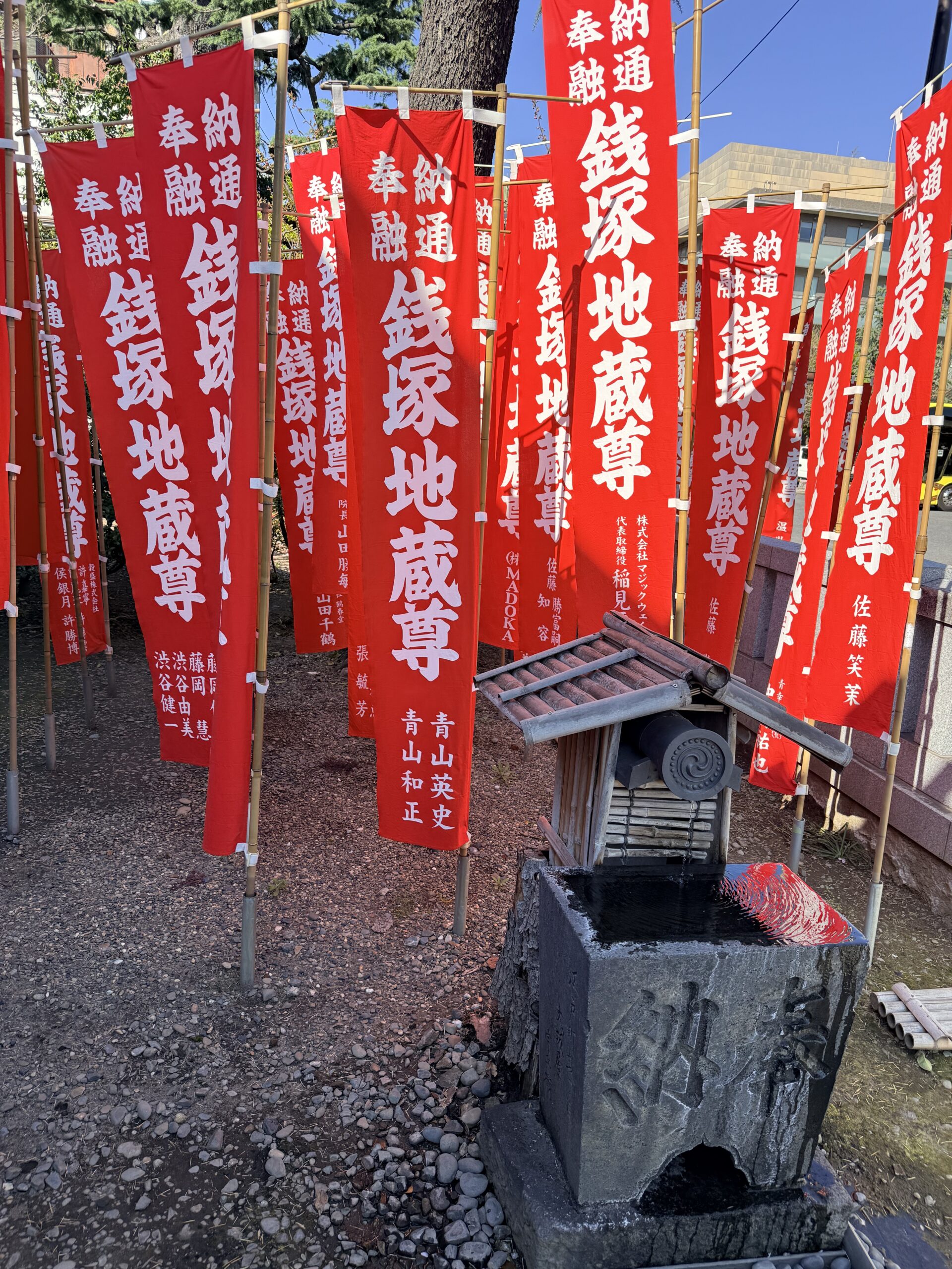 A small stone water basin stands in front of several upright red banners with Japanese writing at a shrine, under bright sunlight.
