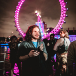 A man stands in front of the illuminated London Eye at night, surrounded by people at an outdoor event.