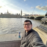 A person takes a selfie by the River Thames with the Houses of Parliament, Big Ben, and Westminster Bridge in the background on a partly cloudy day.