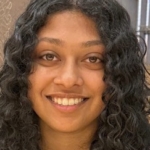 A person with long, curly dark hair and brown skin smiles at the camera in a close-up indoor portrait.
