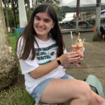 A woman with long brown hair sits outdoors on a concrete ledge, smiling and holding a cup of ice cream. She wears a white T-shirt and denim shorts. Trees and a building are in the background.