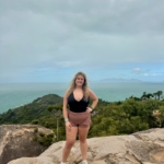 A woman stands on a rocky ledge with her hand on her hip, overlooking green hills and the ocean under a cloudy sky.