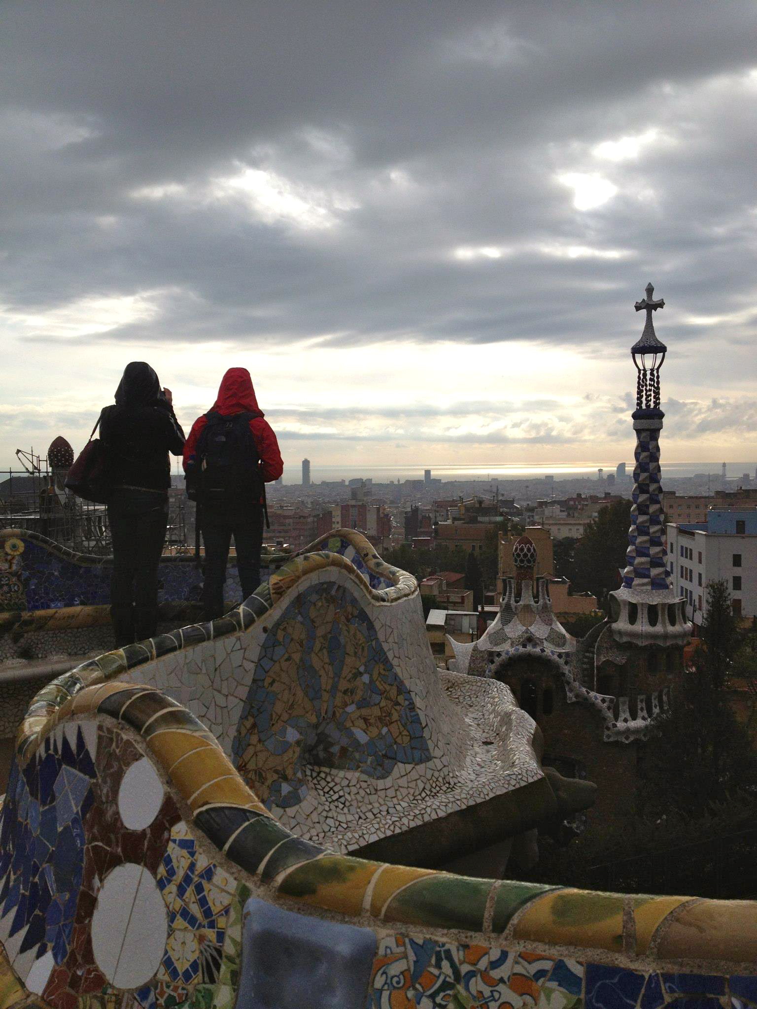 Two people with backpacks stand on a mosaic-tiled terrace overlooking Barcelona's cityscape under a cloudy sky, with Park Güell architecture in the foreground.