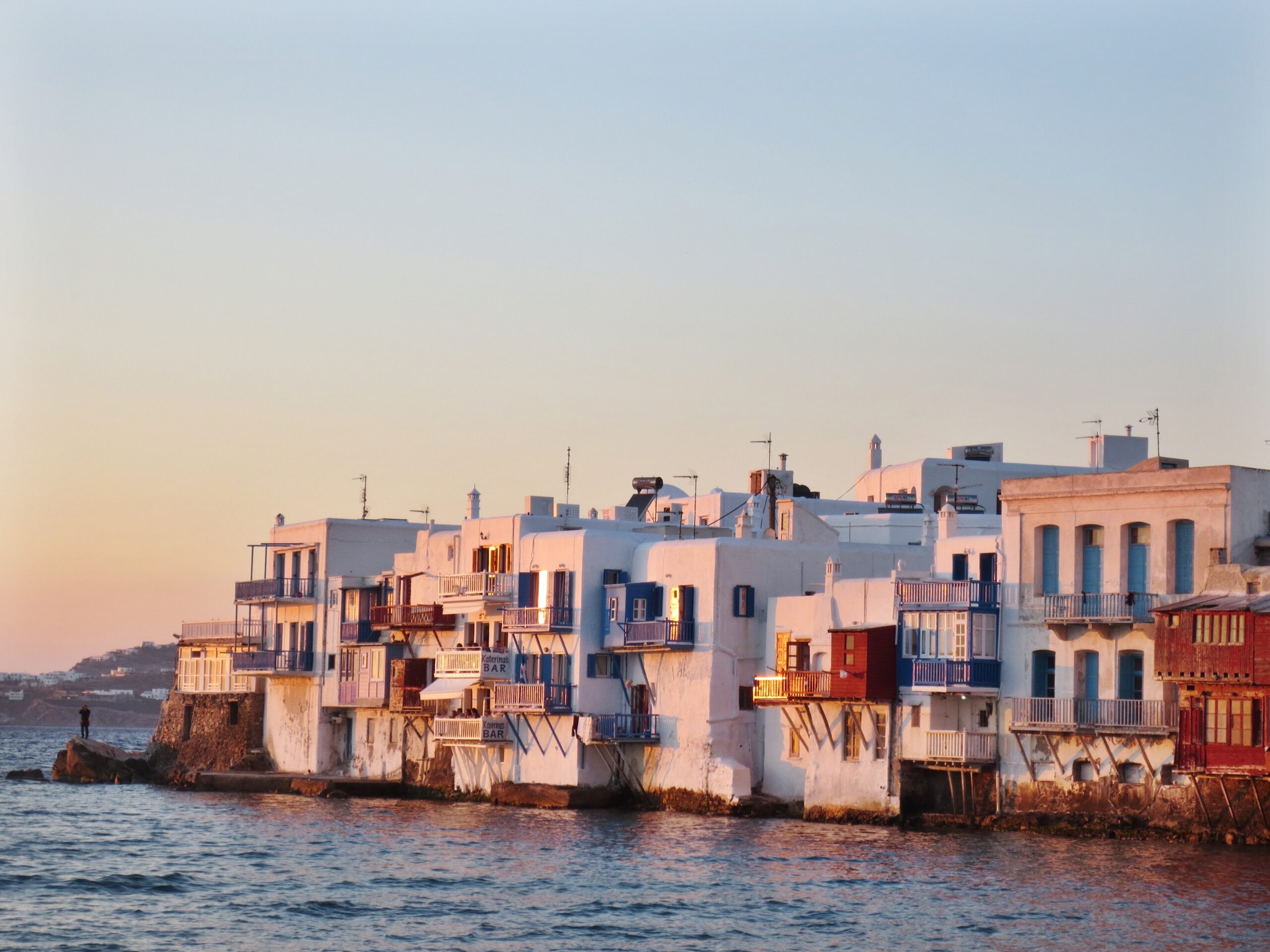 White and blue waterfront buildings stand along the shoreline at sunset, with calm sea water in the foreground.