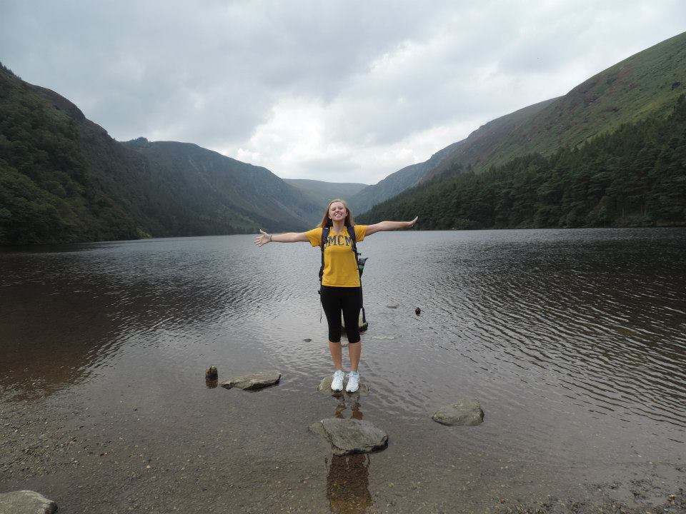 A person stands with arms outstretched on rocks at the edge of a calm lake, surrounded by hills and trees under a cloudy sky.