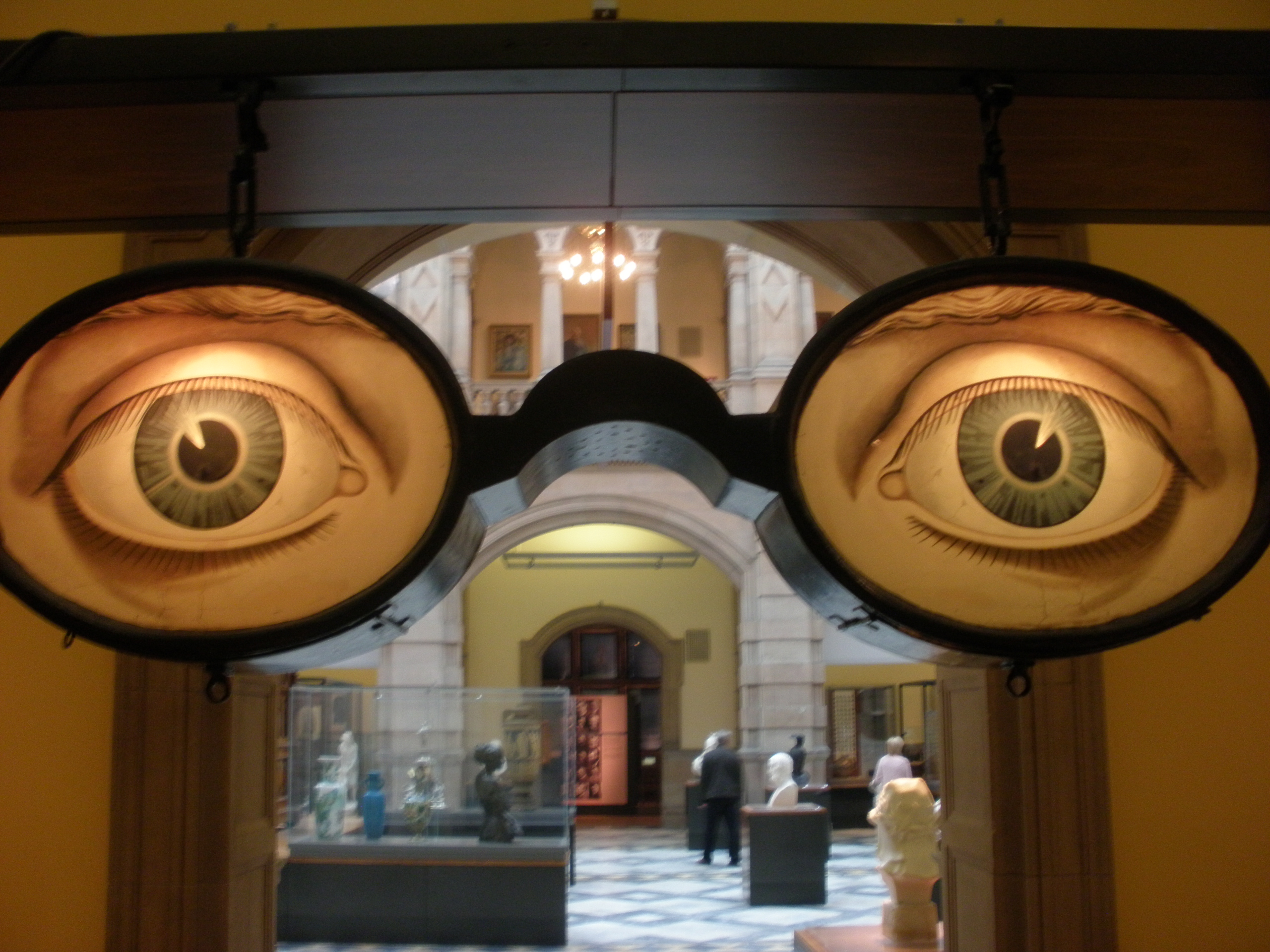 Large sculpture of eyeglasses with illustrated eyes hangs in a museum hallway, with statues and visitors visible in the background.