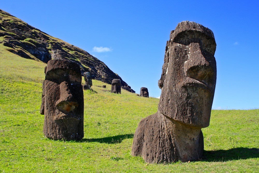 Two large stone Moai statues partially buried in grassy terrain on Easter Island with a blue sky and hillside in the background.