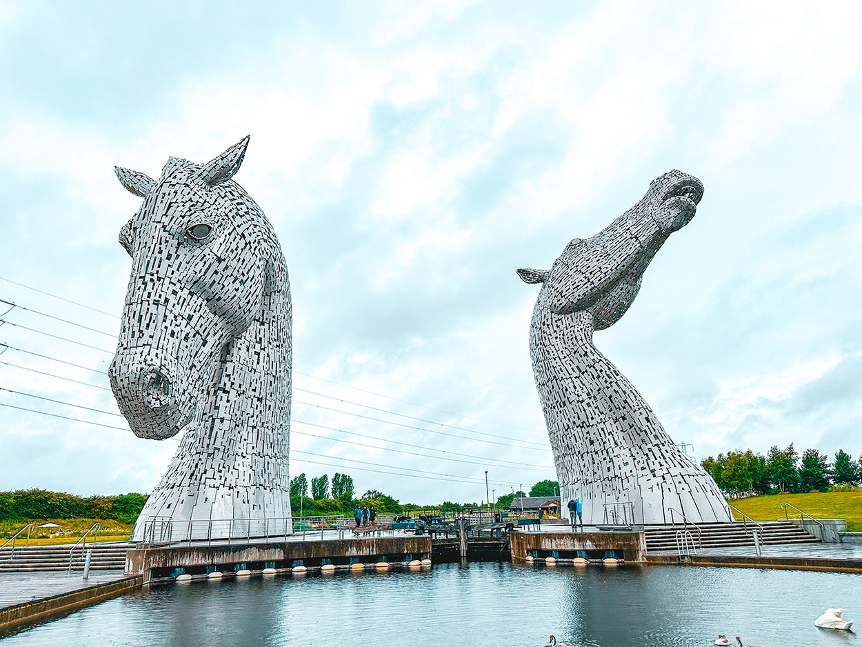 Two large horse head sculptures, known as The Kelpies, stand beside a canal under a cloudy sky with people and greenery in the background.