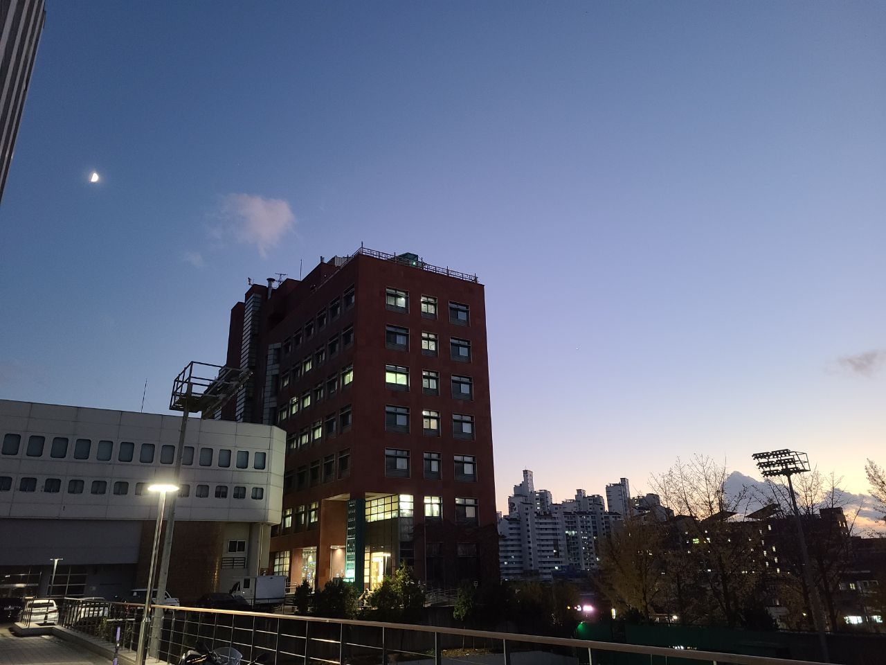 A multi-story brick building stands under a dusk sky with a crescent moon visible; city skyline and streetlights are in the background.