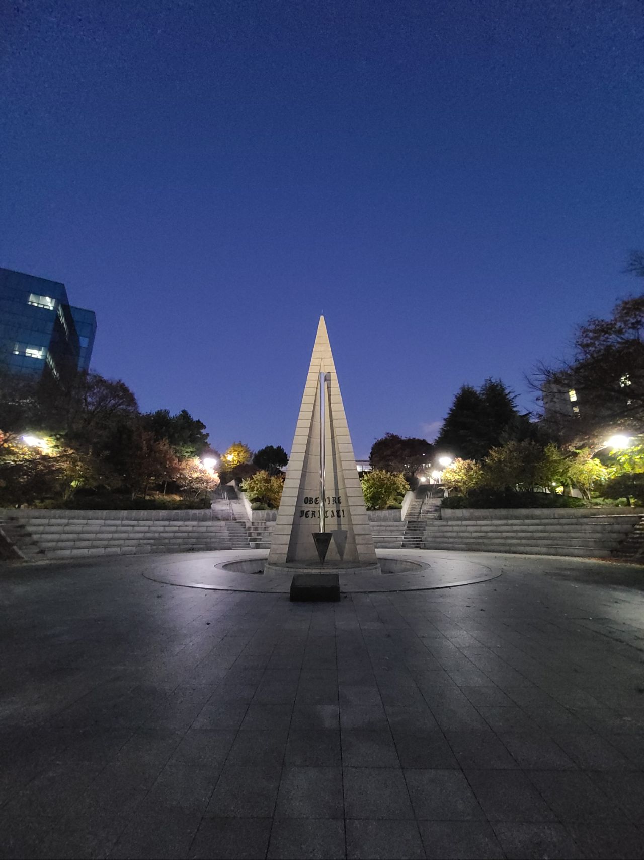 A tall, triangular stone monument stands at the center of a circular plaza, surrounded by illuminated trees and steps, under a clear evening sky.