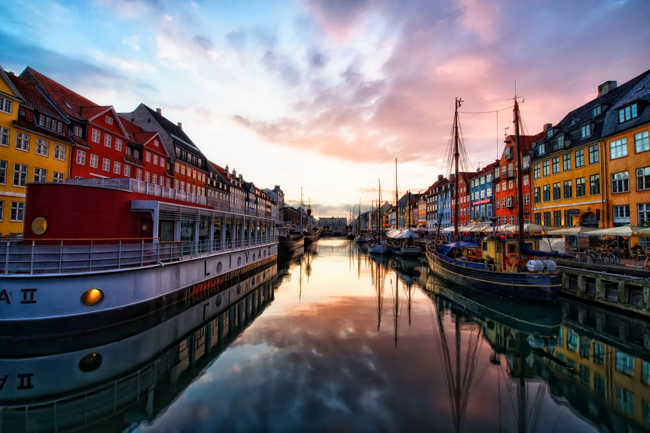 Colorful buildings and boats line both sides of a calm canal at sunset, with a pink and blue sky reflected in the water.