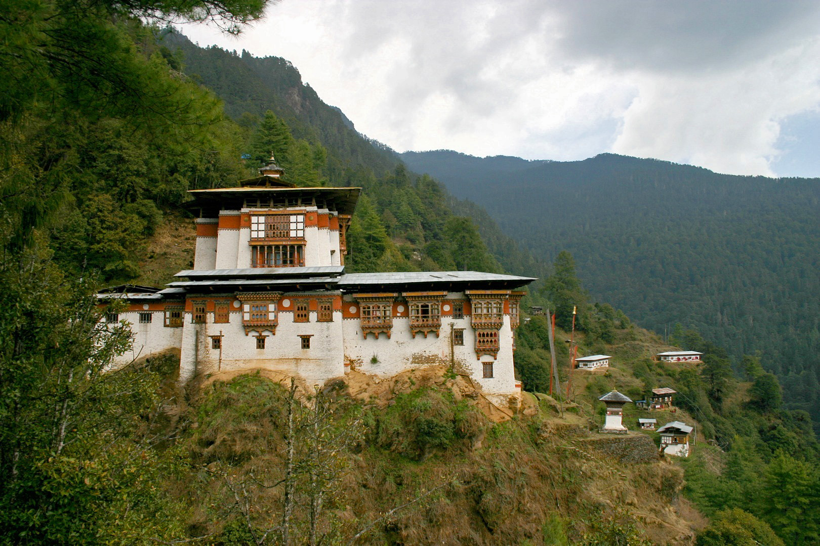 A traditional Bhutanese monastery with white walls and ornate woodwork sits on a forested hillside, surrounded by smaller buildings and green mountains under a cloudy sky.