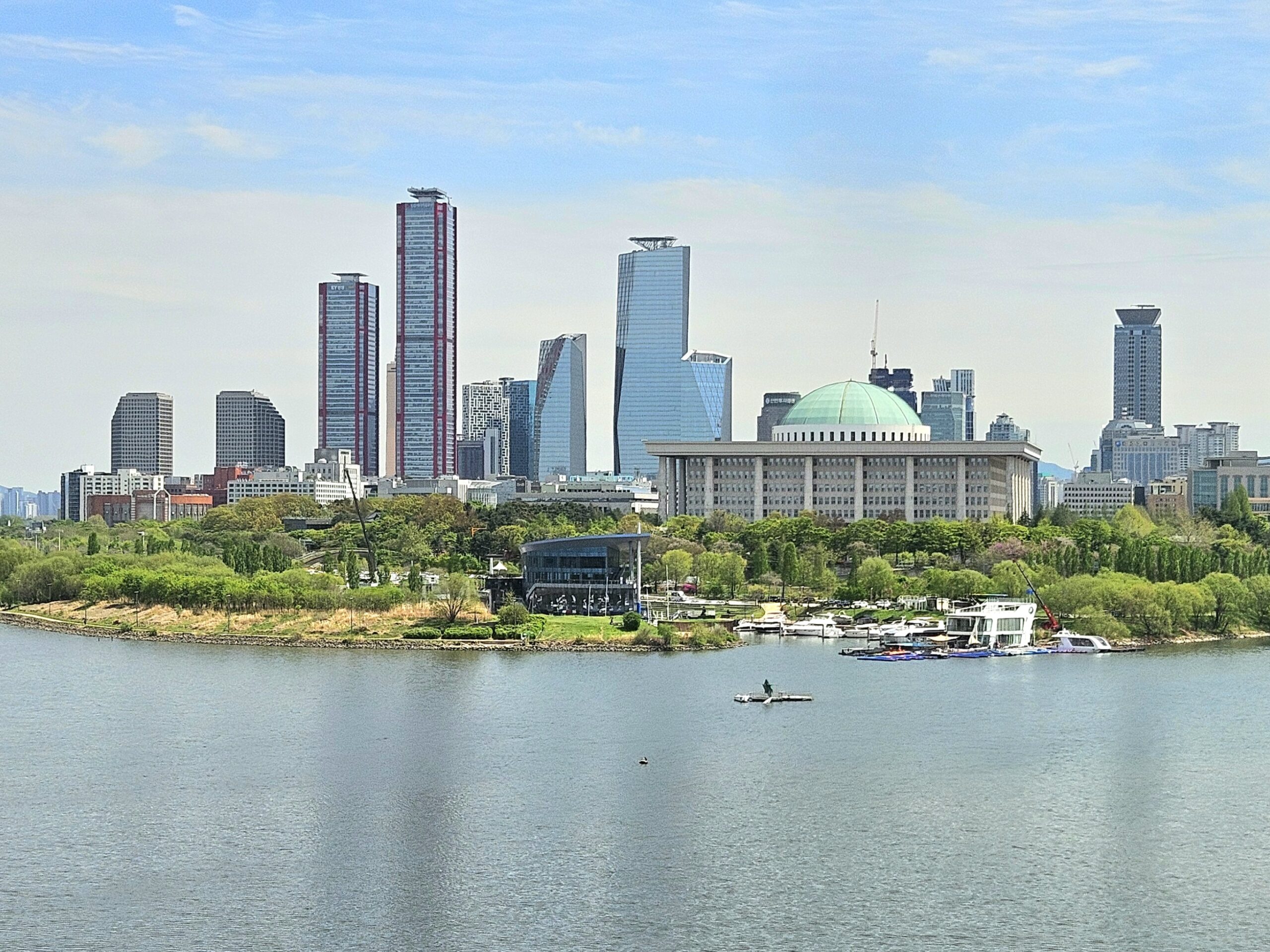 A view of Seoul’s skyline with modern skyscrapers and the National Assembly building, seen from across the Han River under a partly cloudy sky.