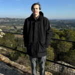 A person wearing a black coat stands in front of a metal railing with a scenic landscape of hills and trees in the background.