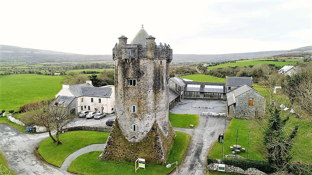 A stone tower stands surrounded by several buildings in a rural landscape with green fields, trees, and distant hills under a cloudy sky.