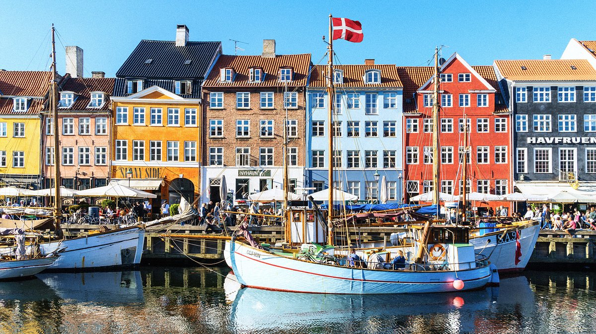 Colorful row houses and boats along the canal in Nyhavn, Copenhagen, with people sitting at outdoor cafes and a Danish flag on one boat.