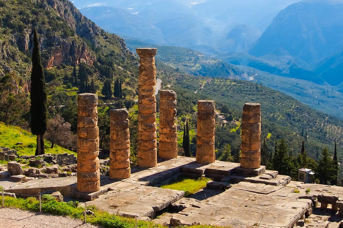 Six ancient stone columns stand amid ruins on a mountain slope, overlooking a green valley and distant hills under a clear sky.