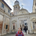 A person stands in front of an old stone archway with a church tower and cross in the background. Other people walk through the arch.