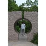 A person stands in the center of a circular stone sculpture outdoors, surrounded by greenery.