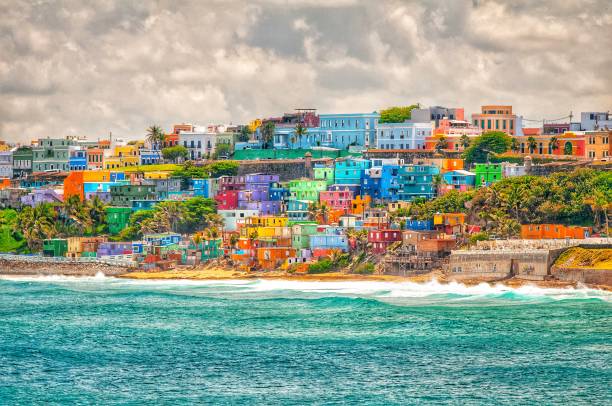 Colorful houses line a hillside by the ocean under a cloudy sky, with waves breaking along the shore in the foreground.