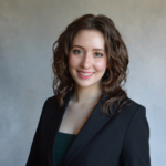 A woman with wavy brown hair, wearing a black blazer over a dark top, smiles at the camera against a neutral gray background.