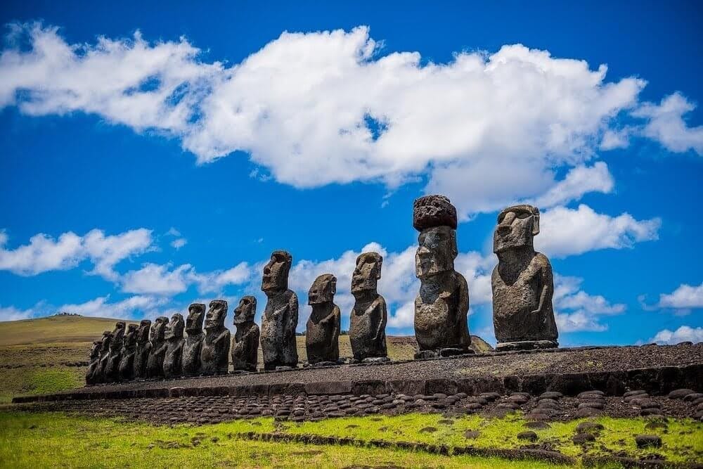 A row of large stone Moai statues stands on a stone platform under a bright blue sky with scattered clouds on Easter Island.