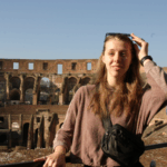 A person stands in front of the Colosseum in Rome, holding sunglasses on their head, with a crossbody bag and long hair.