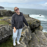 A person wearing sunglasses, a gray top, and jeans stands on rocky cliffs by the ocean under a cloudy sky.