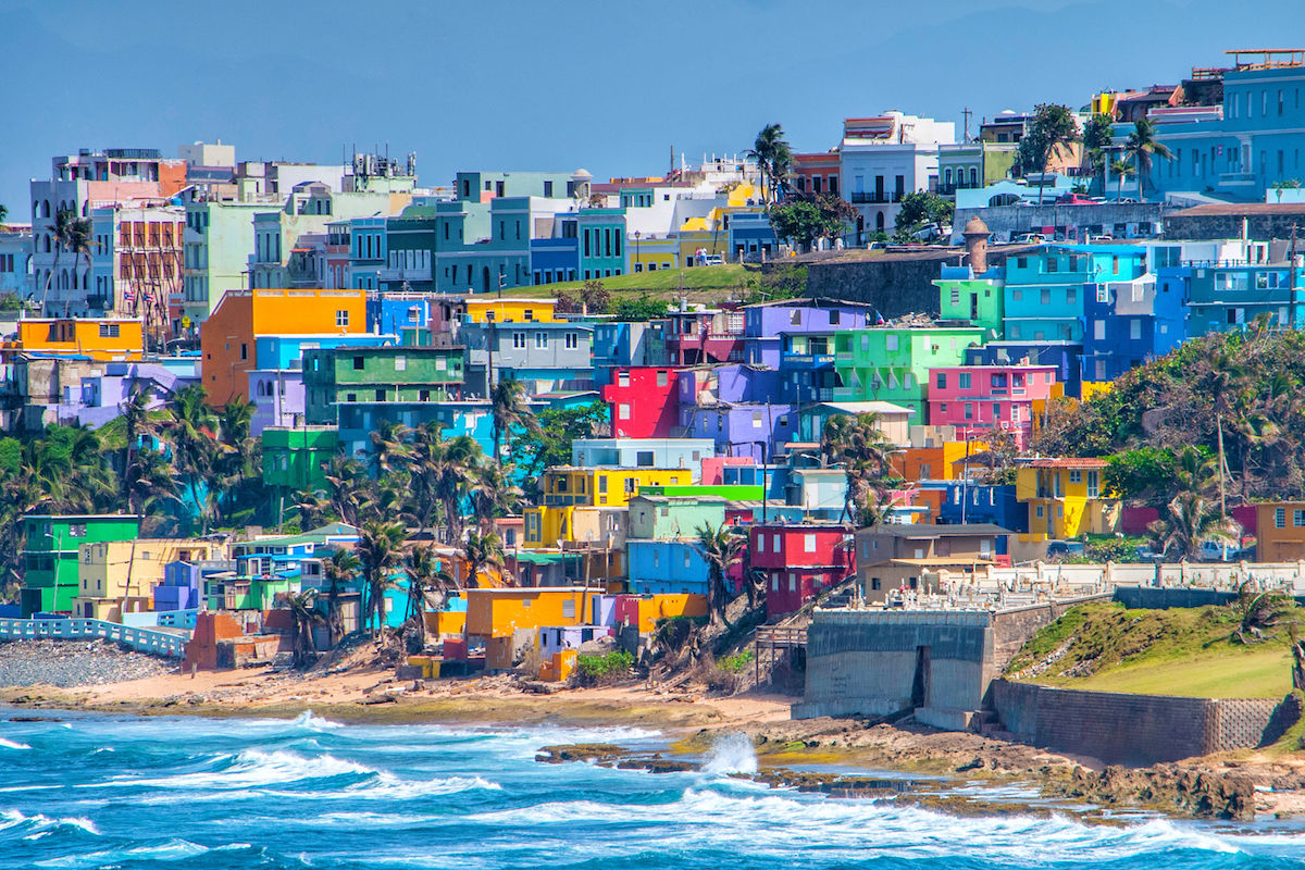 Colorful houses are densely packed on a hillside by the ocean, with waves crashing against the rocky shoreline in the foreground.