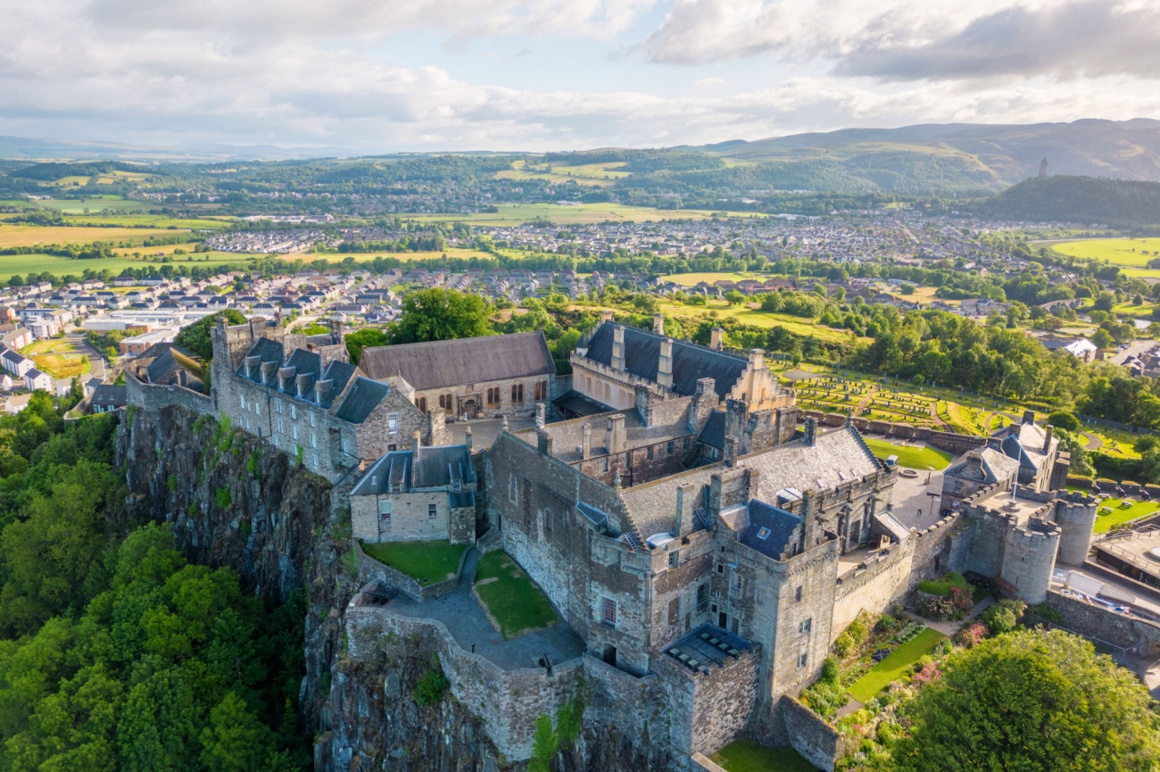 Aerial view of a large stone castle complex on a rocky hill surrounded by greenery, with a town and rolling hills in the background.