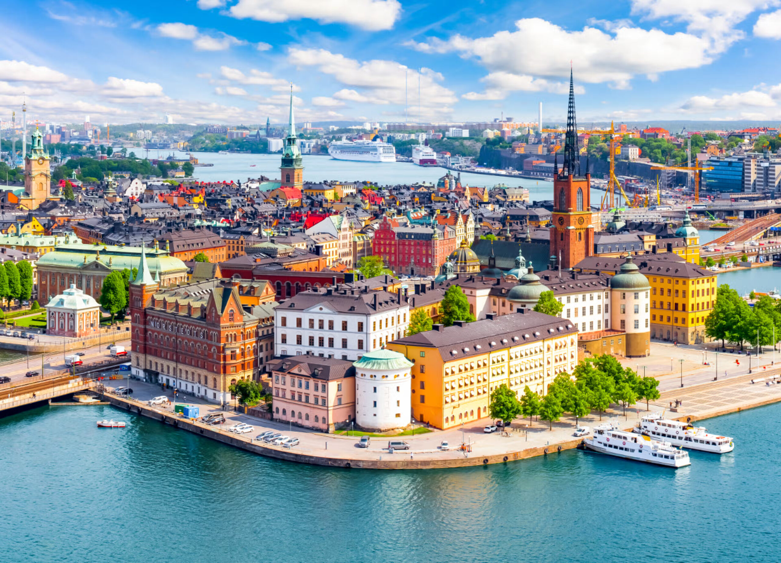 Aerial view of colorful historic buildings, towers, and churches along the waterfront in Stockholm, Sweden, with boats docked and a cityscape in the background.