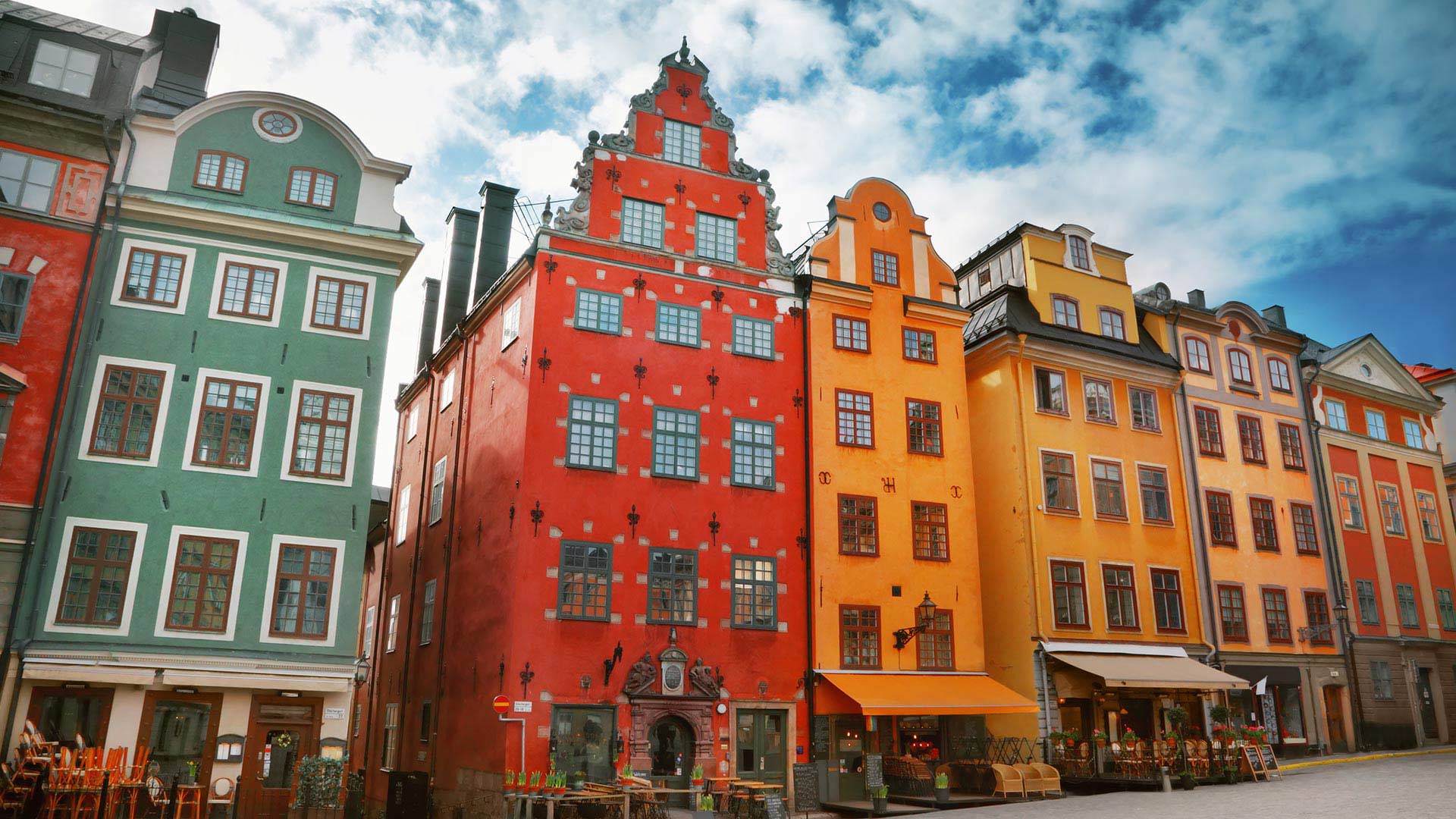 Colorful historic buildings with ornate facades line a cobblestone street under a blue sky with clouds. Several windows and ground-floor shops or cafes are visible.