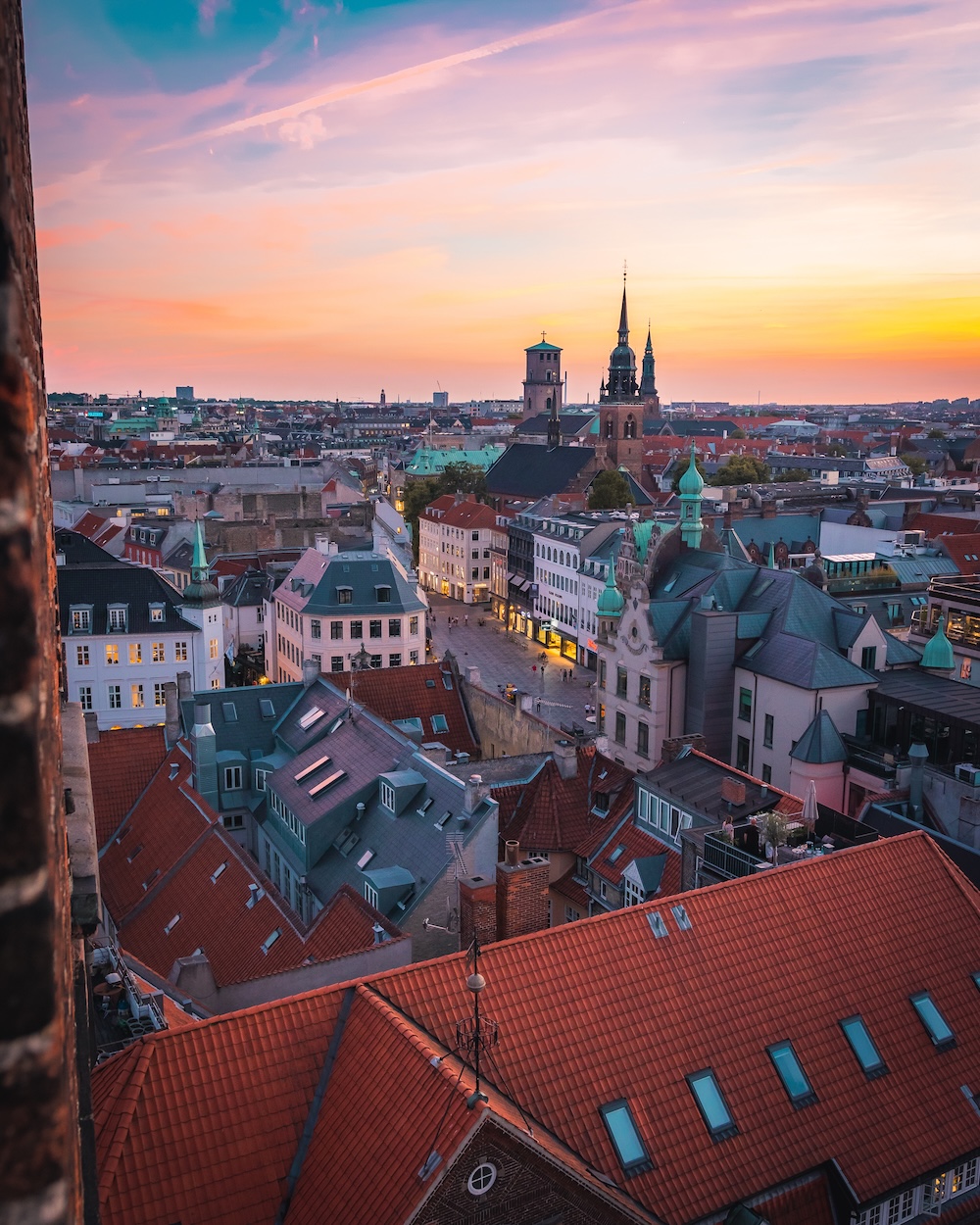 A cityscape at sunset with red-roofed buildings, church spires, and colorful skies over an old European town.