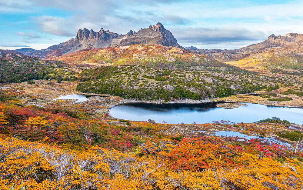 Mountain range with jagged peaks rises behind a lake surrounded by autumn-colored trees and shrubs under a partly cloudy sky.
