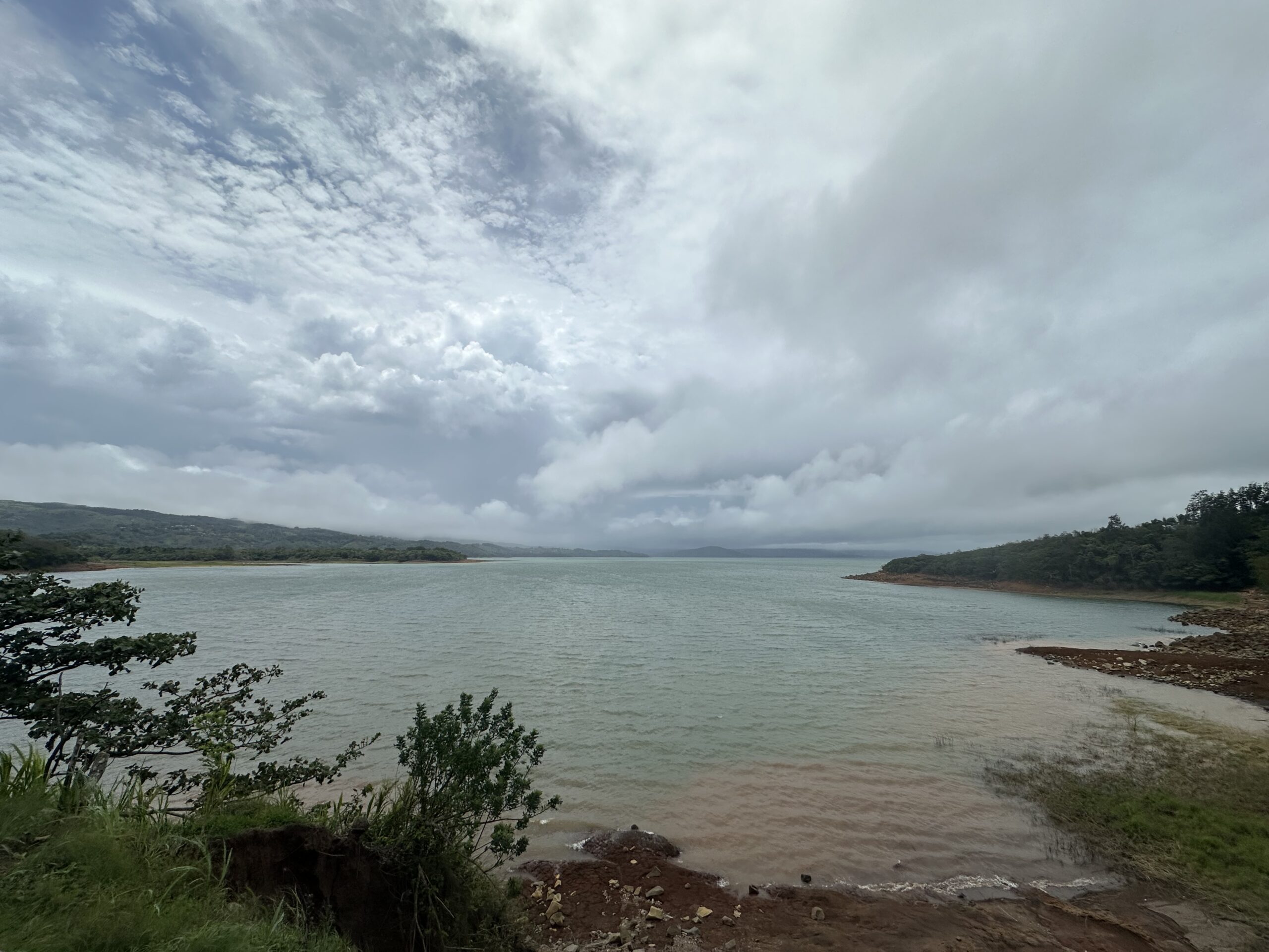 A cloudy sky stretches over a calm lake bordered by trees and reddish-brown soil, with hills visible in the distance.