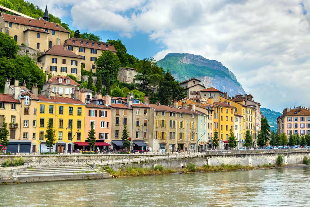 Colorful buildings line a riverbank with a mountain covered in greenery rising in the background under a cloudy sky.