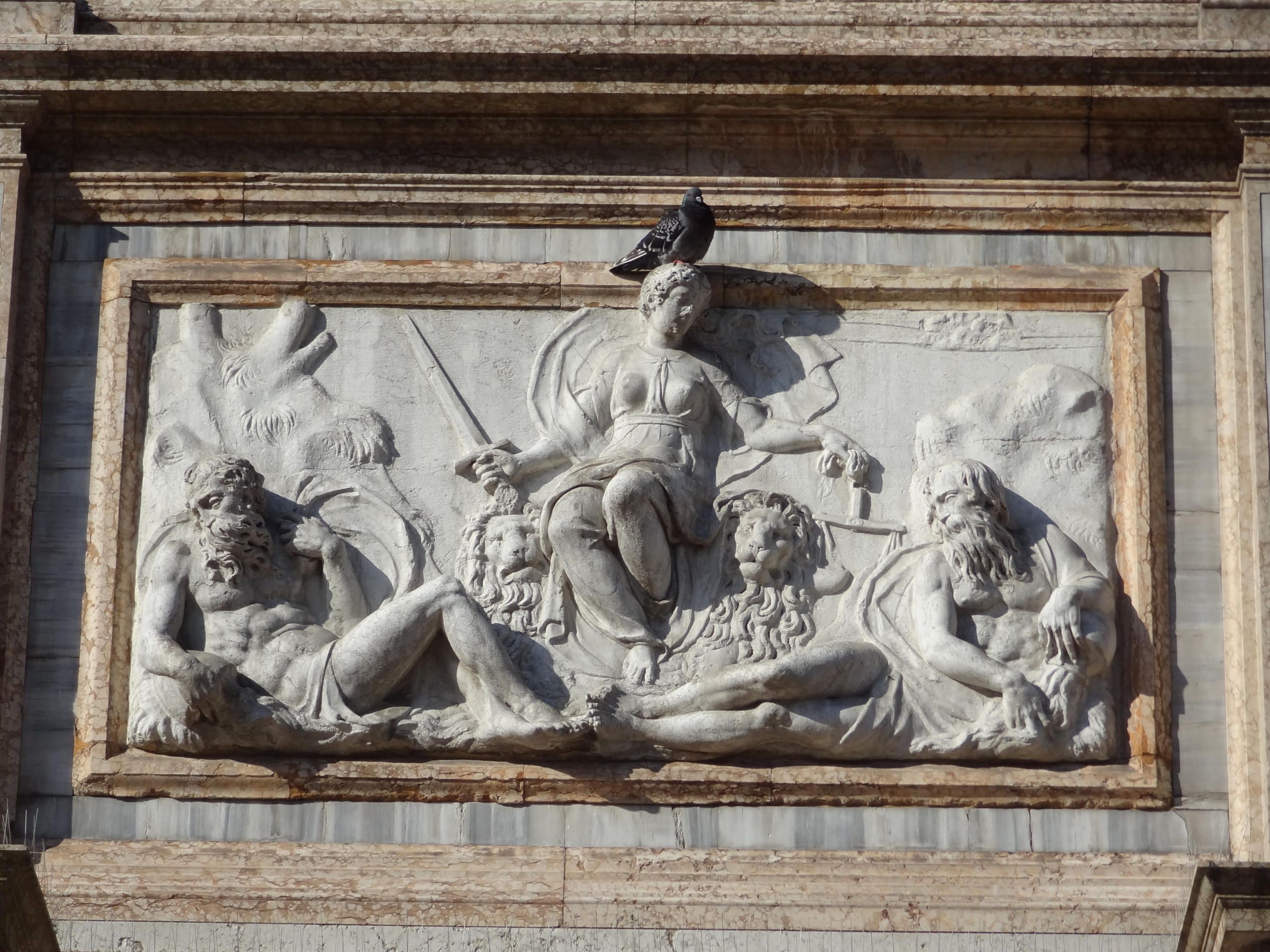 Stone relief sculpture of three figures, with a seated central figure holding a scepter and sword, flanked by two reclining figures; a pigeon is perched on top.