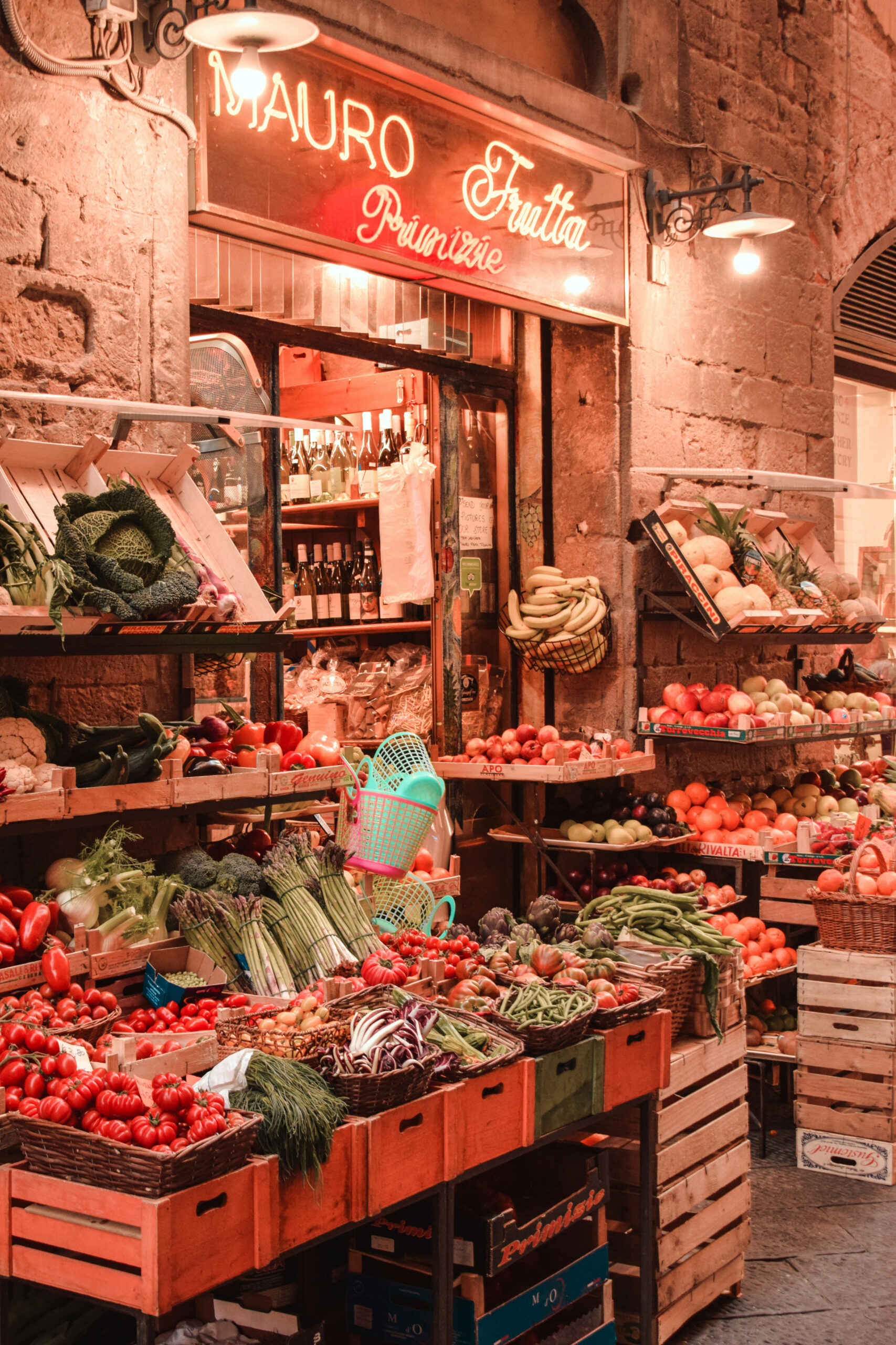 Outdoor fruit and vegetable market stand displays fresh produce in wooden crates under a neon sign and warm lighting in front of a stone building.