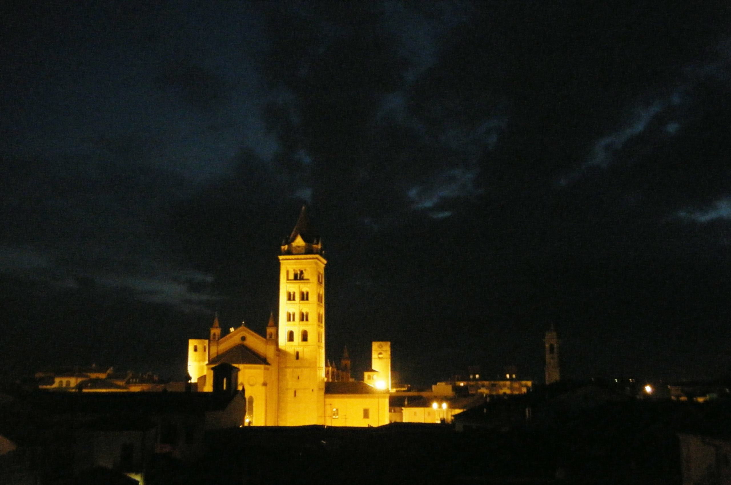 A brightly lit church and bell tower stand out against a dark, cloudy night sky in an urban area.
